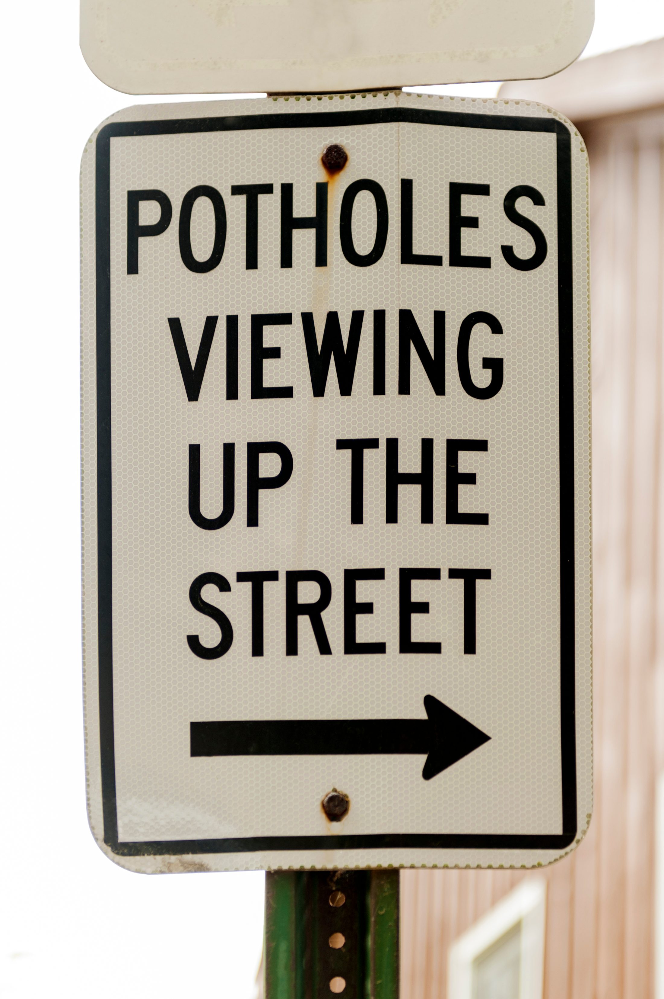 A metal road sign directs viewing up the street, indicating pothole presence. An arrow points right, against a blurred urban background.