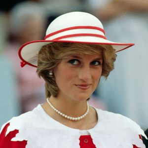 Person wearing a hat and pearl necklace, smiling subtly, surrounded by blurred crowd in a formal outdoor setting.