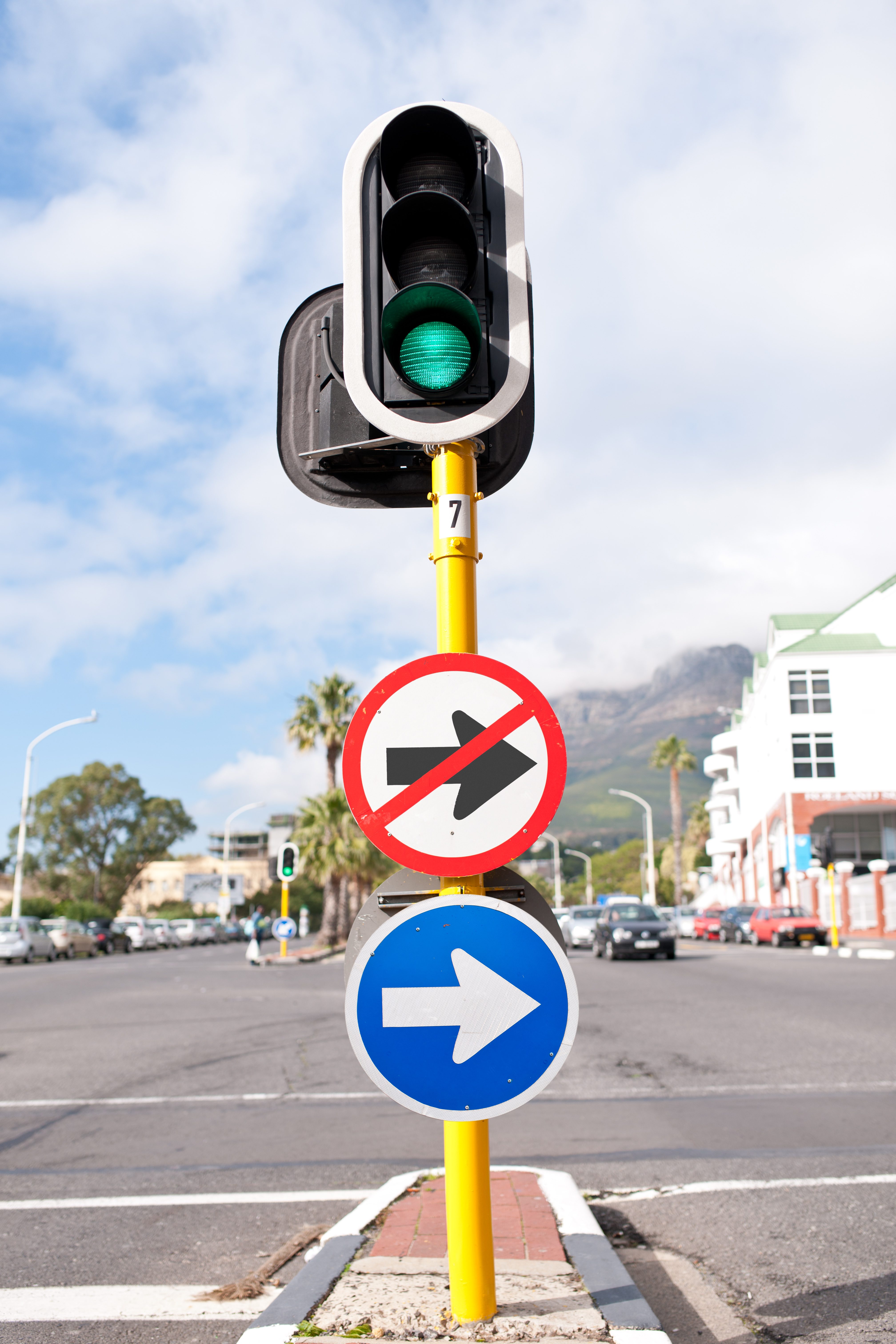 Traffic light shows green, with signs indicating no right turn and mandatory right. Urban street context with cars, buildings, and mountains in the background.