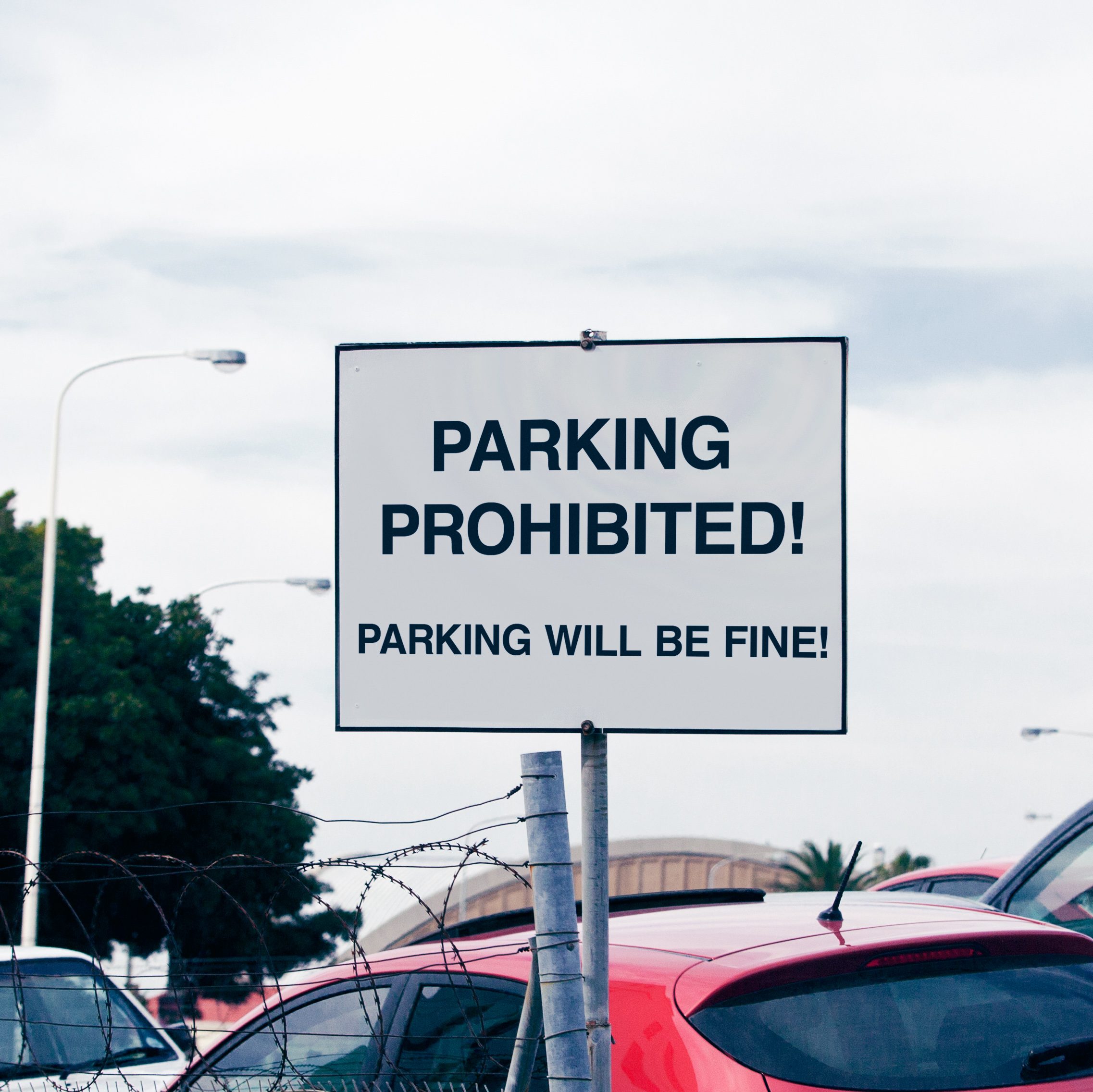 Sign displays conflicting messages, "PARKING PROHIBITED!" and "PARKING WILL BE FINE!" over parked cars, surrounded by barbed wire and trees.