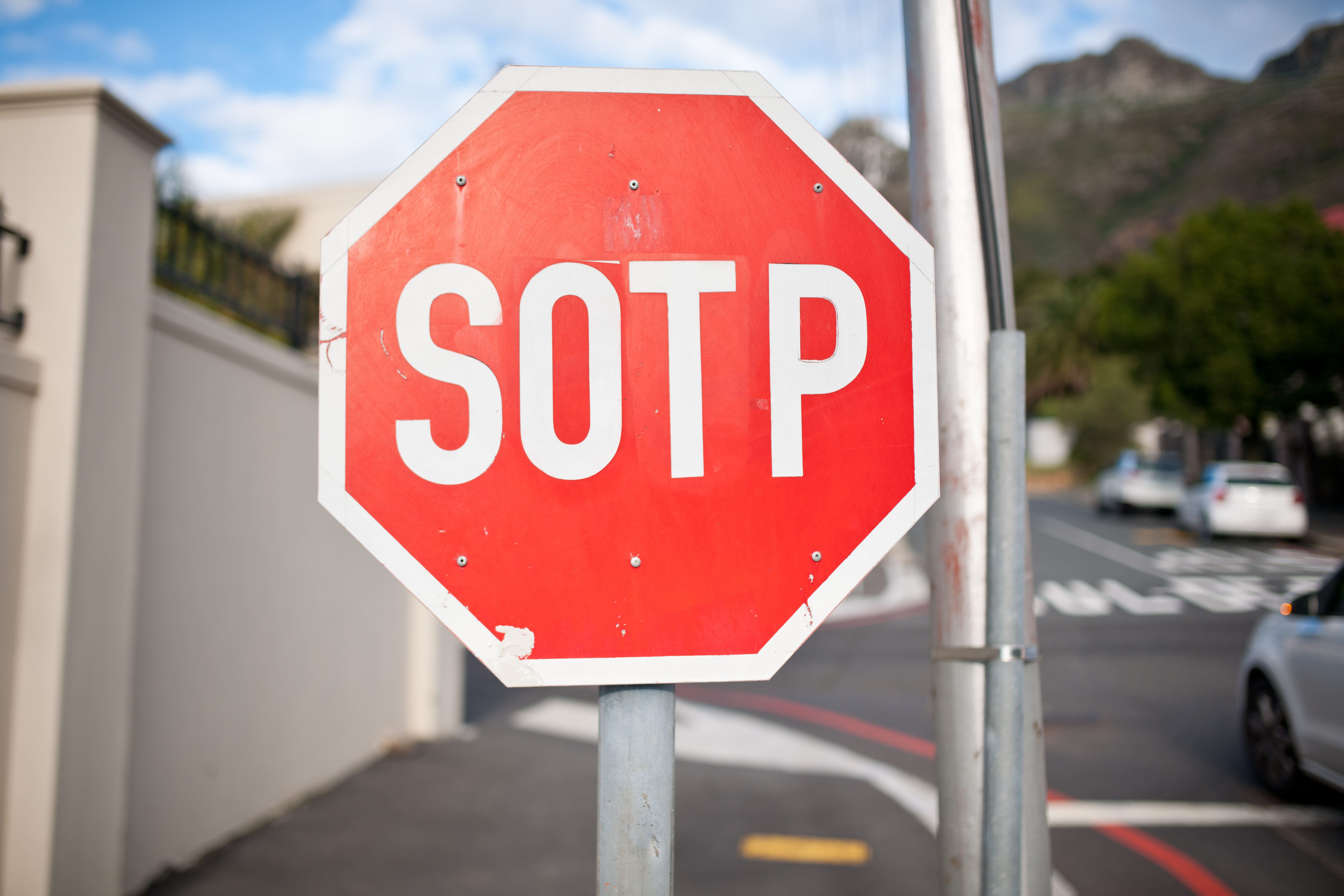 Red octagonal sign displays "SOTP," positioned at roadside intersection beside a white wall and scenic background with mountains and trees.