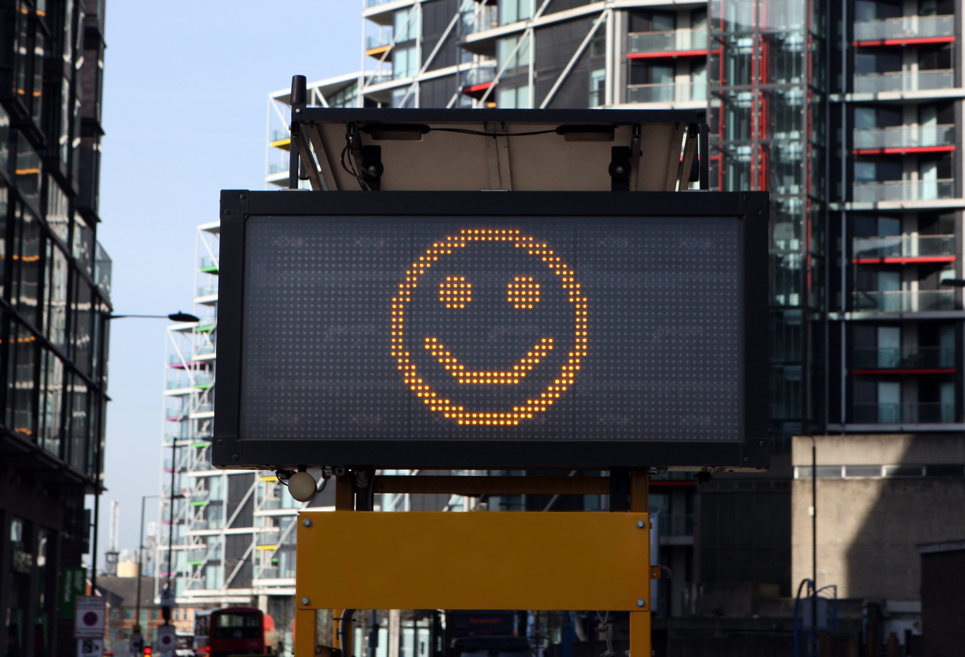 Digital sign displays a yellow smiling face, standing amidst tall urban buildings on a city street.