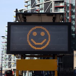 Digital sign displays a yellow smiling face, standing amidst tall urban buildings on a city street.