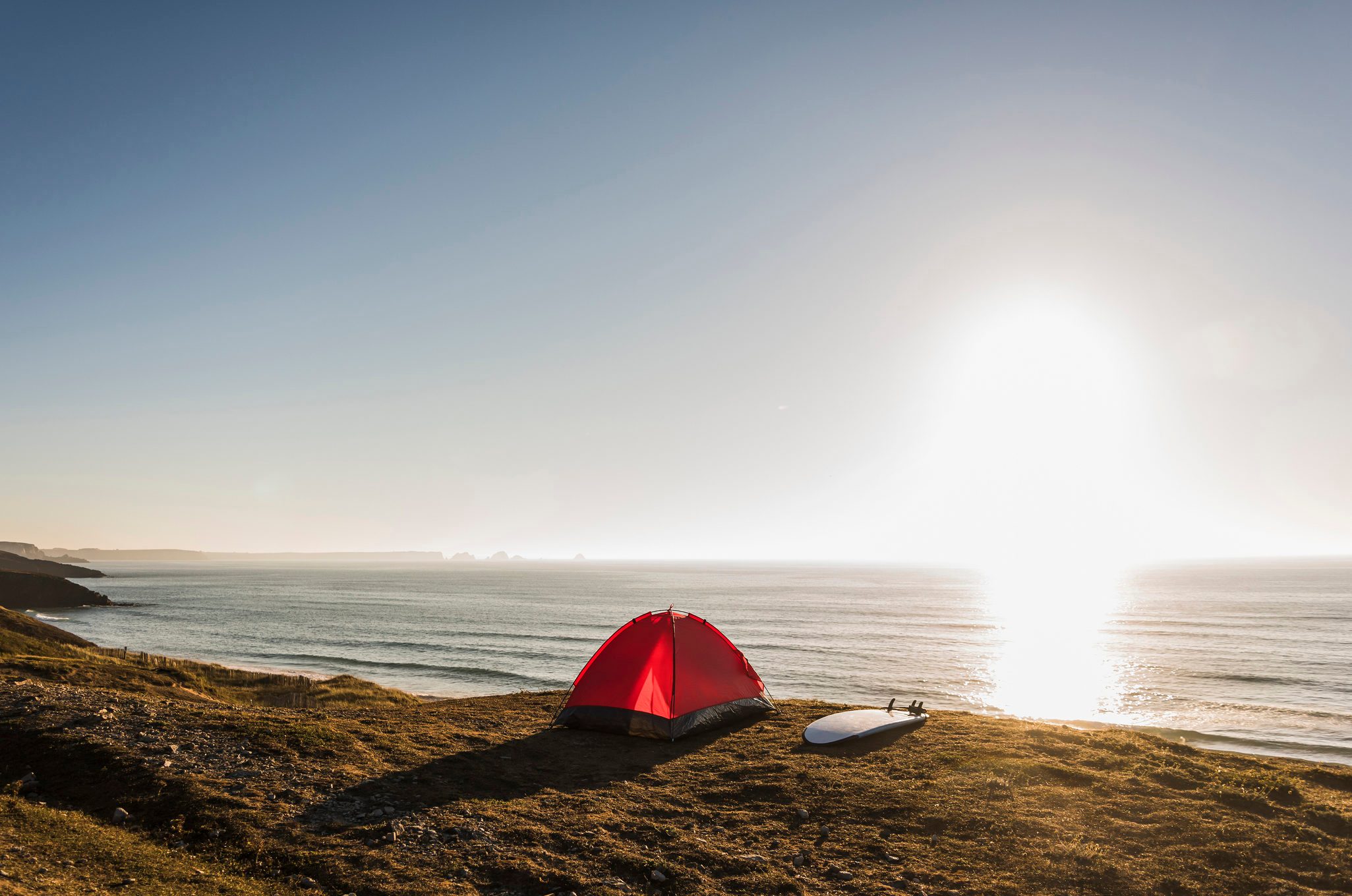A red tent stands still beside a surfboard on a grassy cliff overlooking the ocean during a bright sunset.