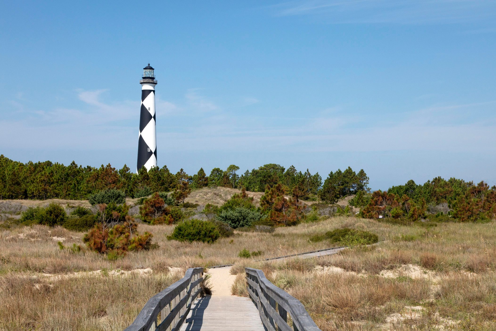 Lighthouse stands tall, surrounded by grass and trees, under a clear blue sky, with a wooden path leading through sandy terrain.