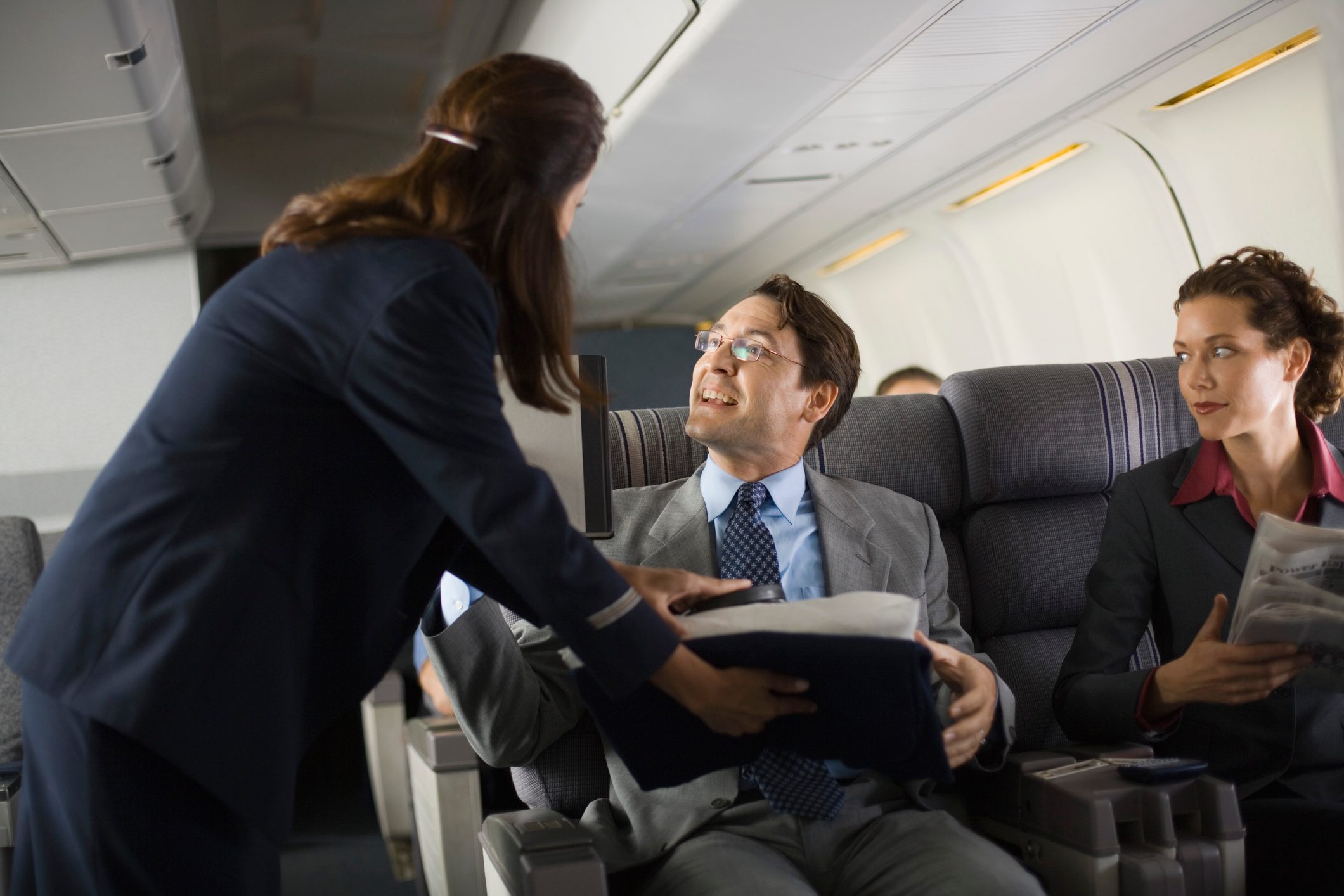 Flight attendant hands pillow to seated man smiling, woman holding newspaper looks on, inside airplane cabin.
