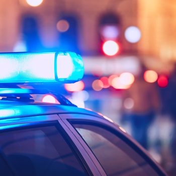 Police car emits blue light; city street blurred with people and glowing red lights in background.
