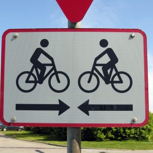 Sign depicts cyclists moving left and right, with arrows indicating directions, set against a clear sky and green surroundings.
