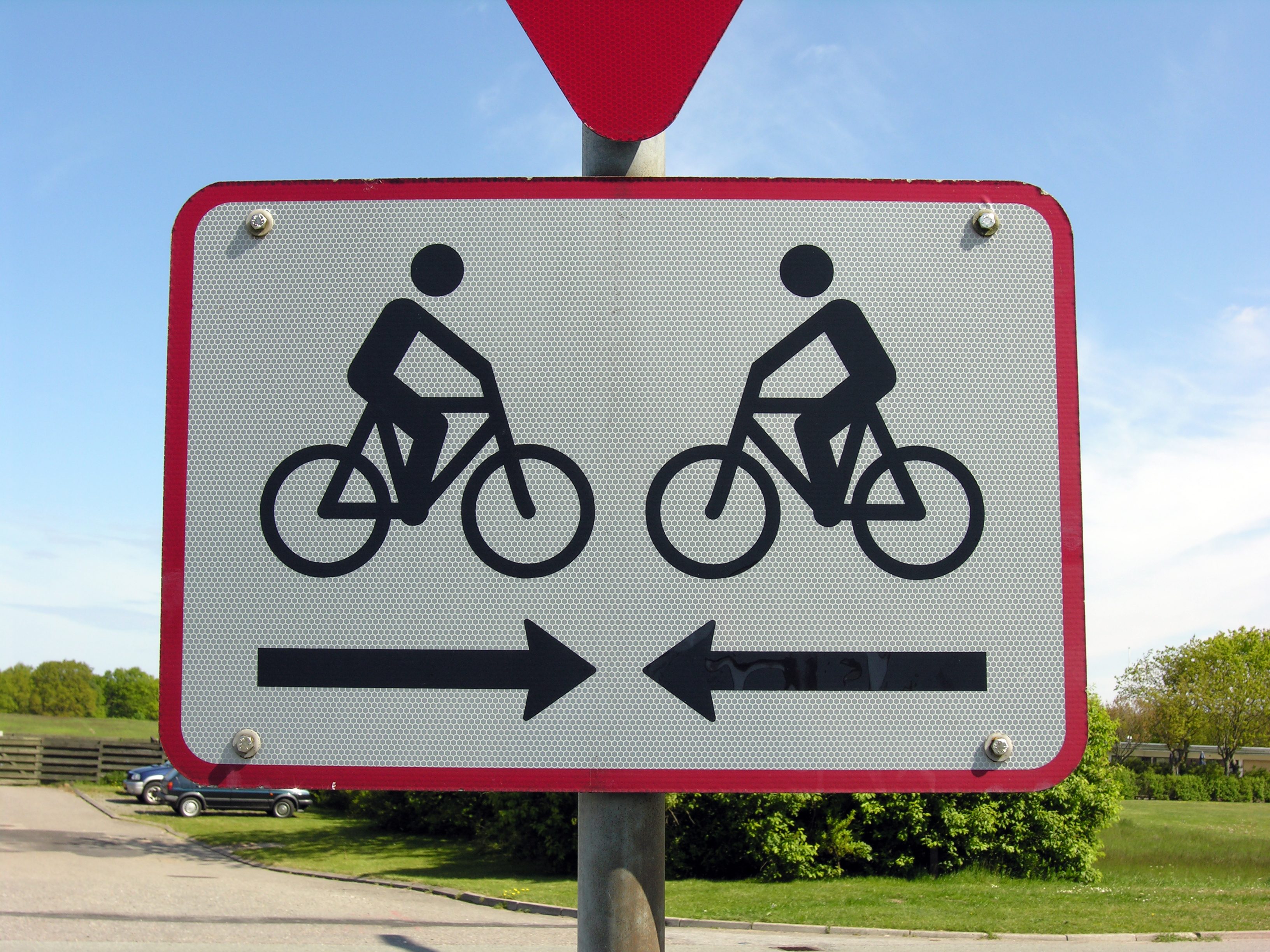 Sign depicts cyclists moving in opposite directions, accompanied by arrows pointing left and right, with a backdrop of greenery and sky.