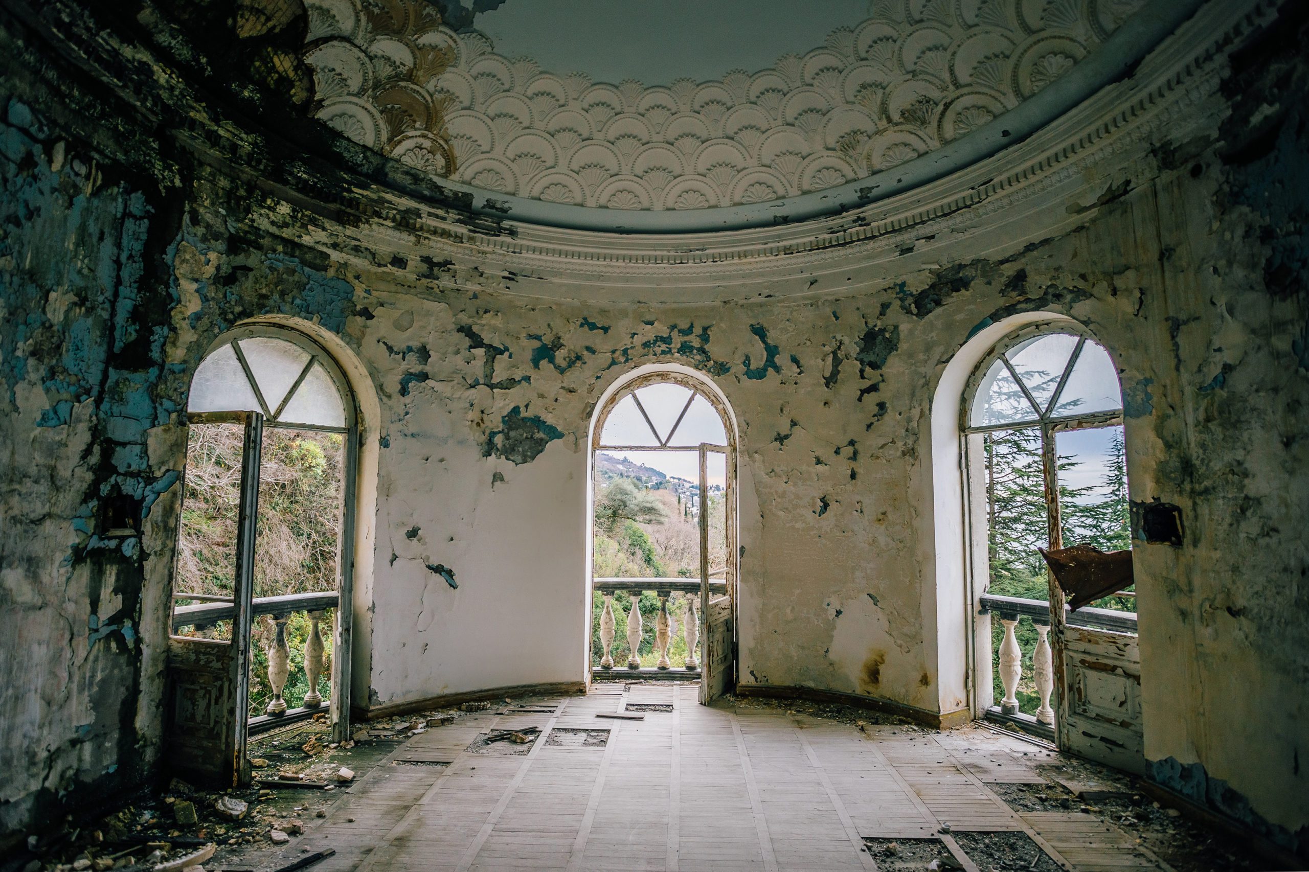 Room with peeling walls and broken windows overlooks lush, hilly landscape. Dusty, decayed interior contrasts with vibrant greenery outside, suggesting abandonment.