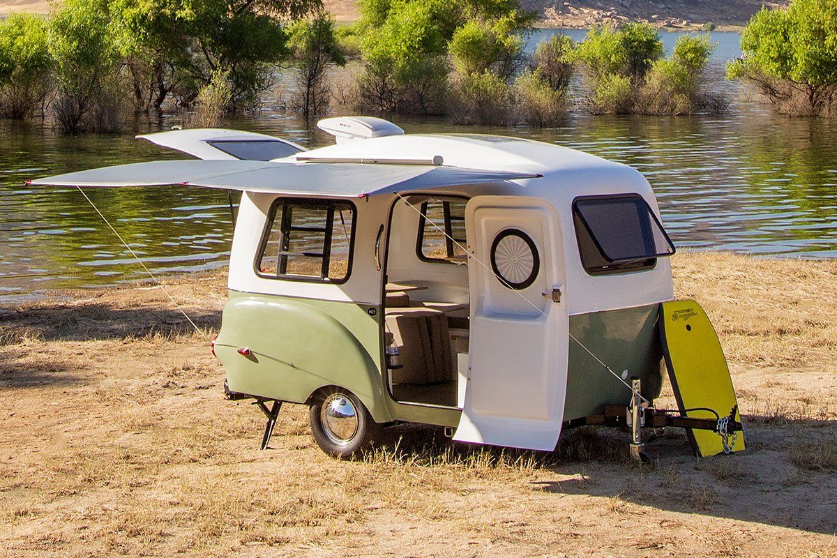 Camper trailer stands with door open, displaying interior, parked on grassy lakeside, surrounded by green trees and calm water.