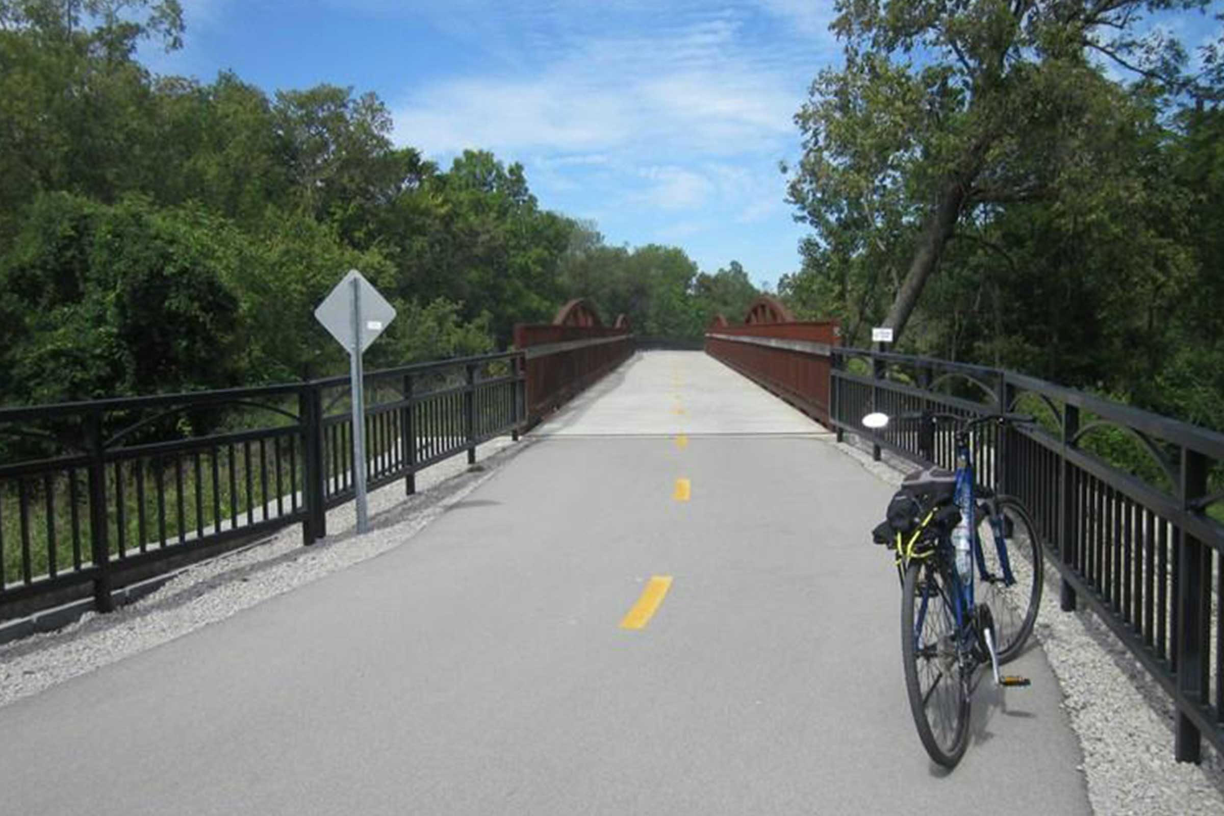 Bicycle rests on bridge, surrounded by lush green trees under a clear blue sky.