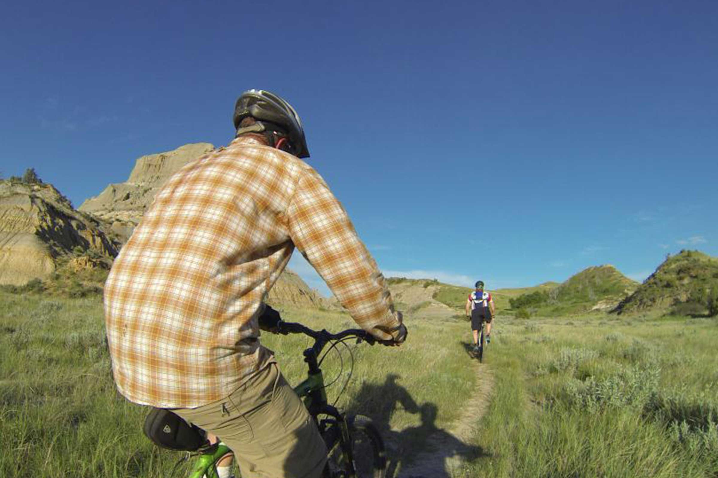 Cyclist rides along grassy trail, surrounded by hills under a clear blue sky, followed closely by another cyclist in similar terrain.