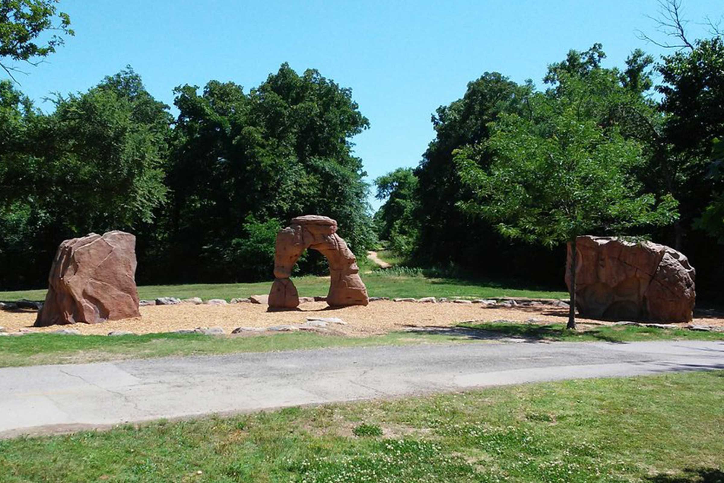 Rocks stand arranged in an open, grassy park with a path and dense trees in the background under a clear blue sky.
