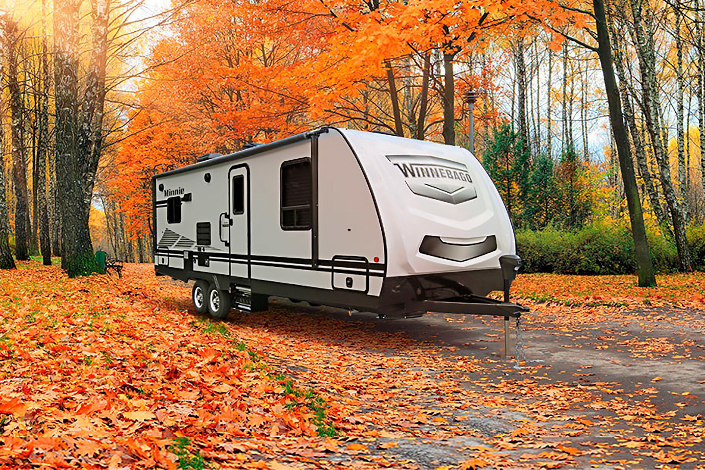 Trailer stationed in vibrant autumn forest, surrounded by fallen orange leaves. Text on trailer reads "Winnebago" and "Minnie."