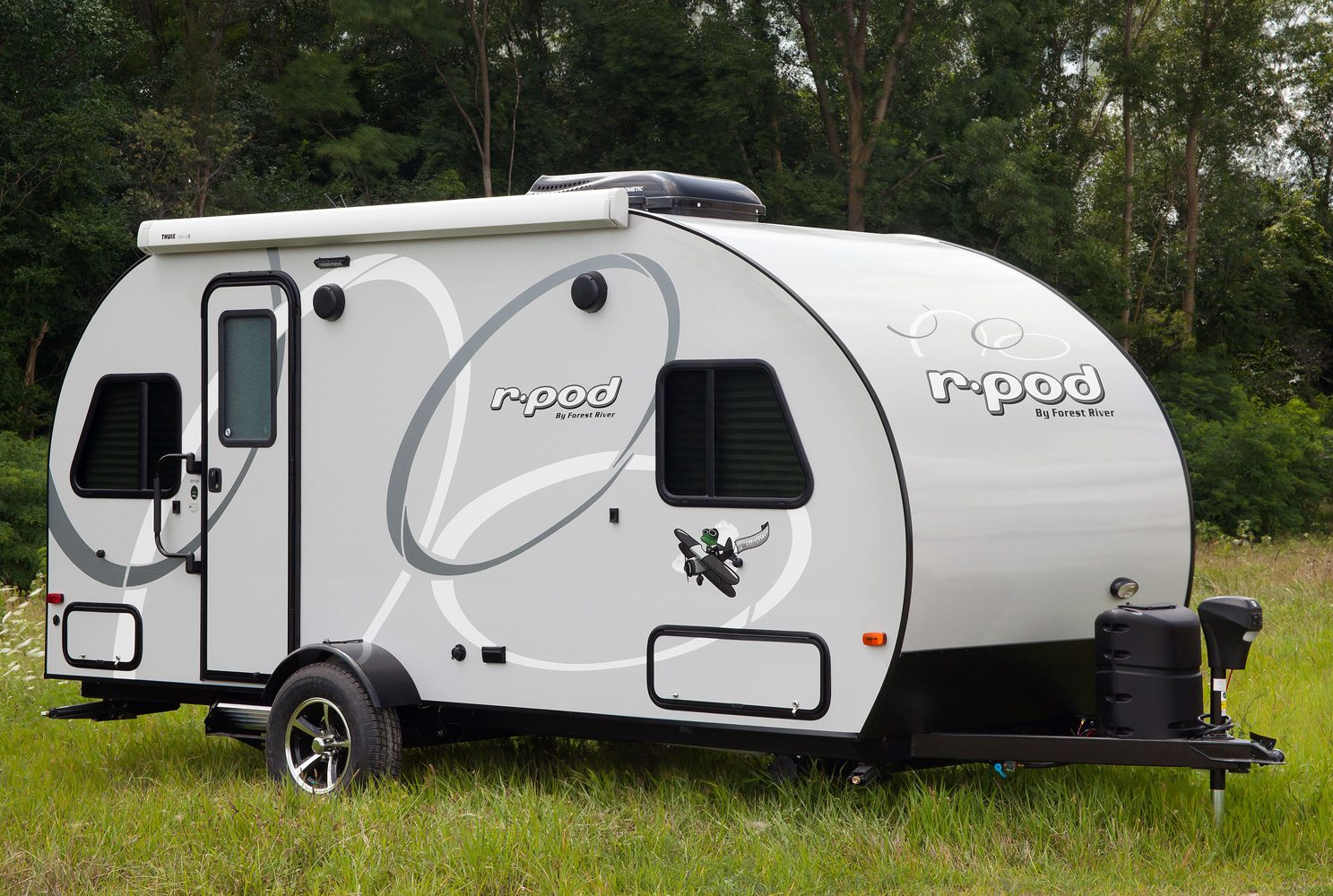Trailer resting on grass, featuring rounded edges with "râ€¢pod by Forest River" text on the side; surrounded by dense forest.