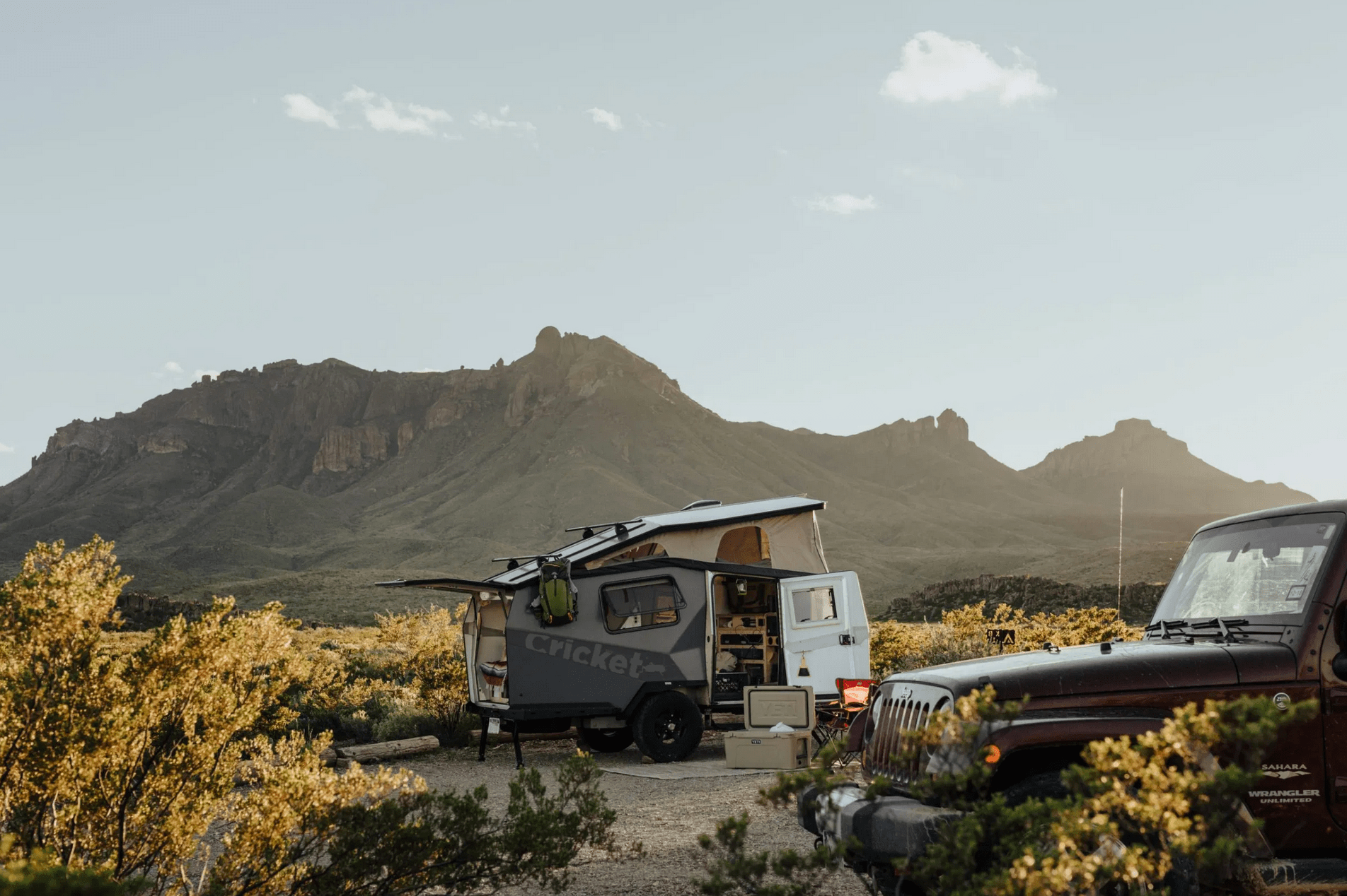 Camper parked with an open door in a desert landscape, adjacent to an SUV, under a clear sky with mountains in the background.