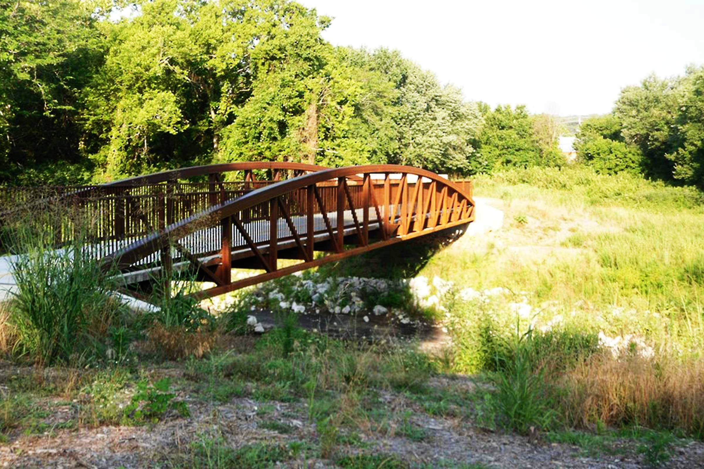 A wooden footbridge spans over a small stream, surrounded by lush greenery and tall grass under a clear sky.