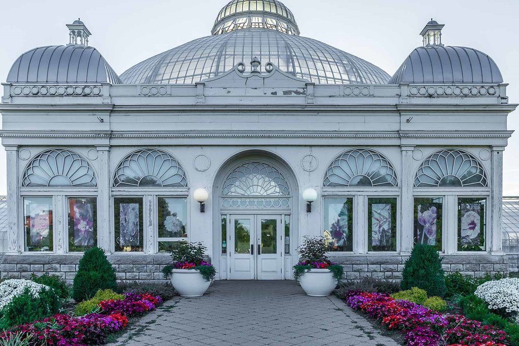 A glass-domed conservatory stands surrounded by vibrant flowerbeds, showcasing an elegant architectural design with arched windows and intricate detailing.