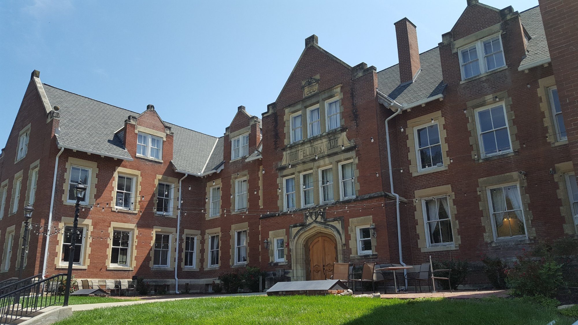 Brick building stands in sunlight, featuring gothic architectural details and green lawn; string lights hang across facade, enhancing its historic appearance.