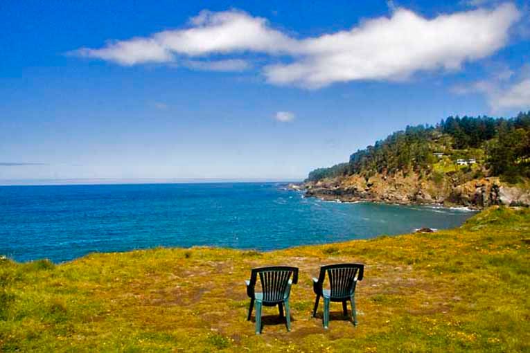 Two empty chairs face the ocean, overlooking a grassy cliff with a rocky coastline and a bright, cloudy sky.