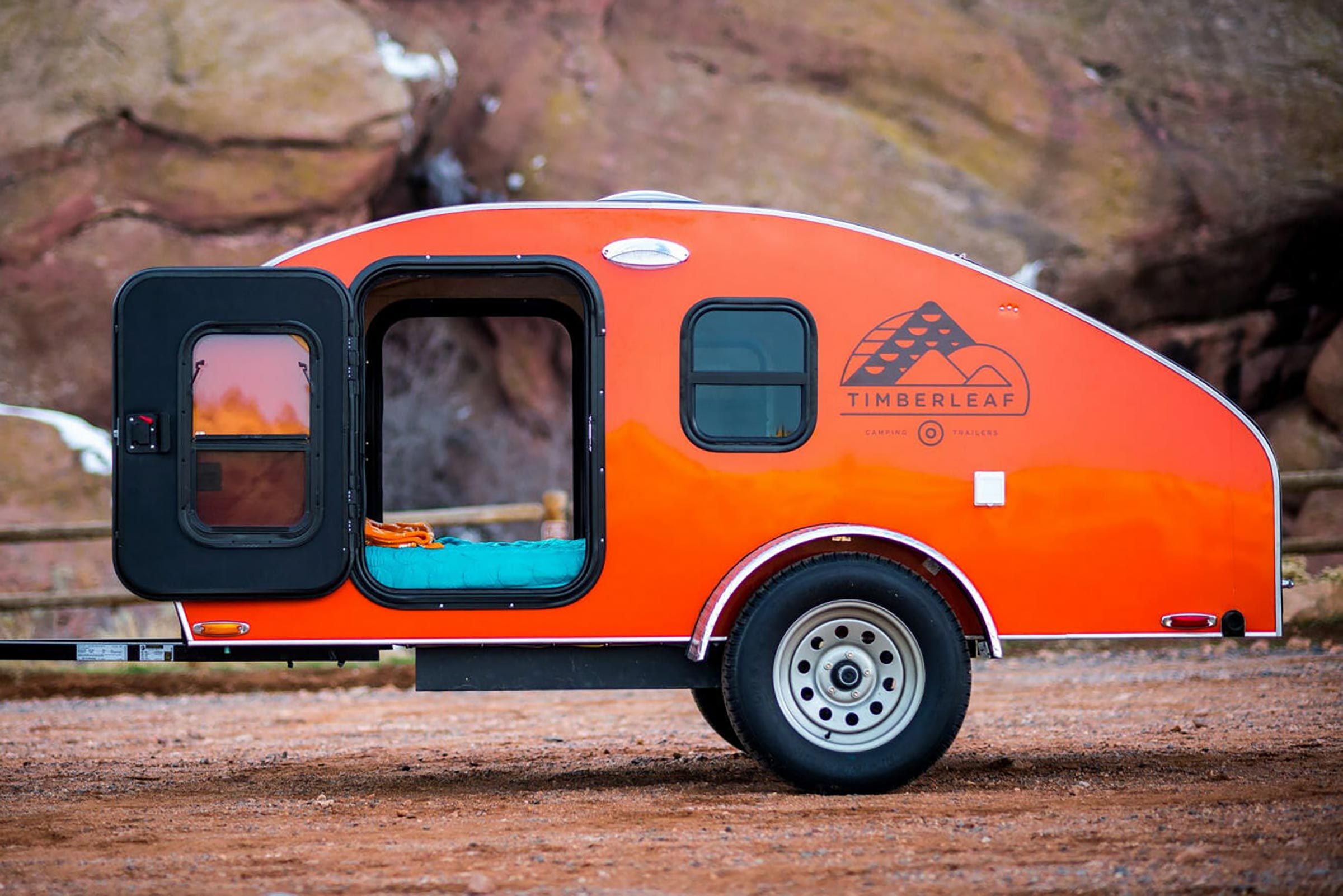 Trailer with open door, revealing a cozy interior, parked on rocky terrain. The trailer is bright orange with "Timberleaf" logo on the side.