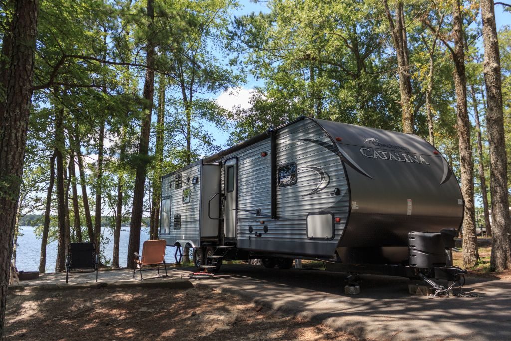 Camper parked in wooded area near a lake, with chairs outside. Text reads "Catalina" on camper side.