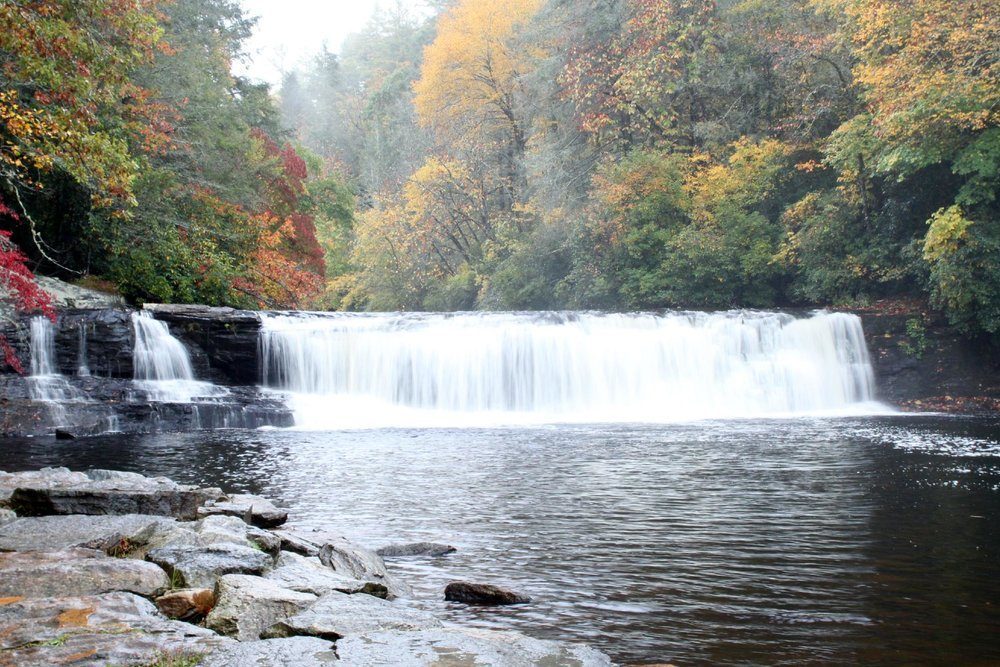 A waterfall cascades gently over rocks, surrounded by vibrant autumn trees with colorful foliage, into a calm, reflective pool.