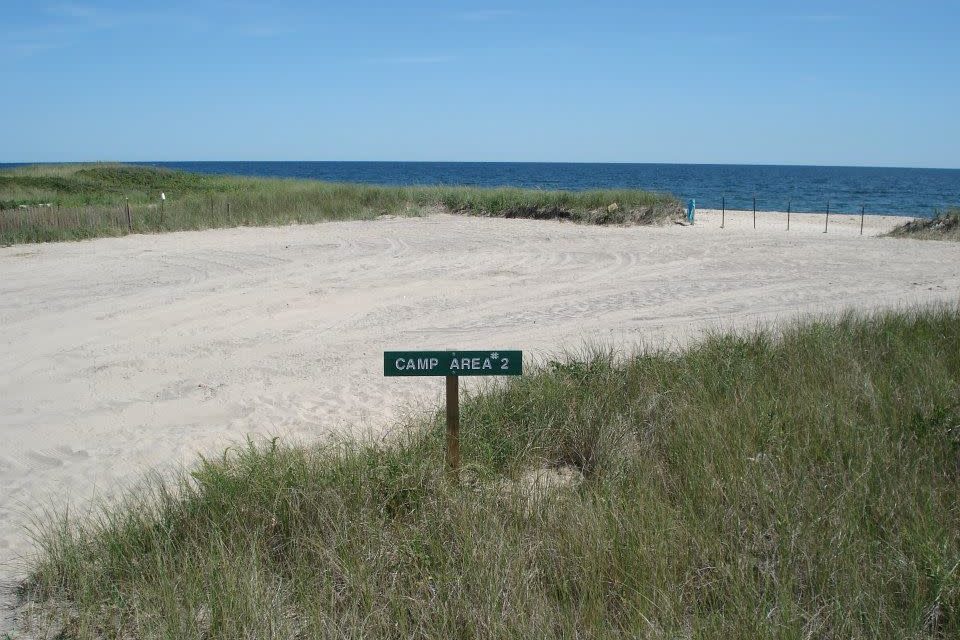 Sign reading "CAMP AREA 2" stands amid grassy dunes near a sandy beach, overlooking the blue ocean under a clear sky.