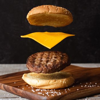 Burger ingredients float above a wooden board: sesame bun, cheese slice, patty, and bottom bun. Salt scattered around. Dark background.