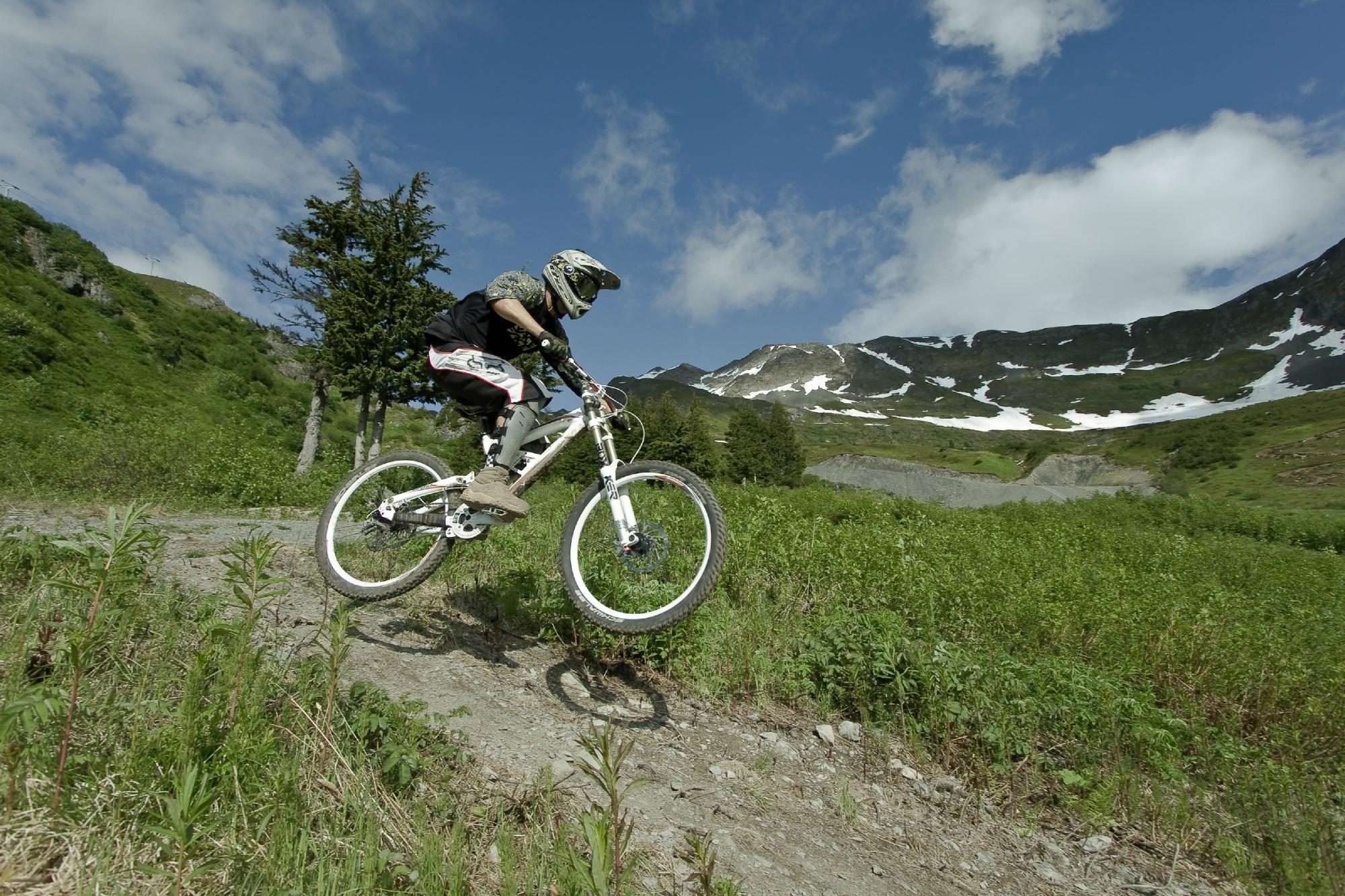 Cyclist jumps on a mountain bike along a dirt trail, surrounded by greenery and snow-capped mountains under a blue sky.