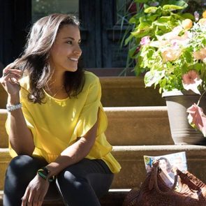 Woman sits smiling on steps, playing with hair. She's wearing a yellow blouse, surrounded by potted flowers and a woven bag. Sunlight illuminates her.