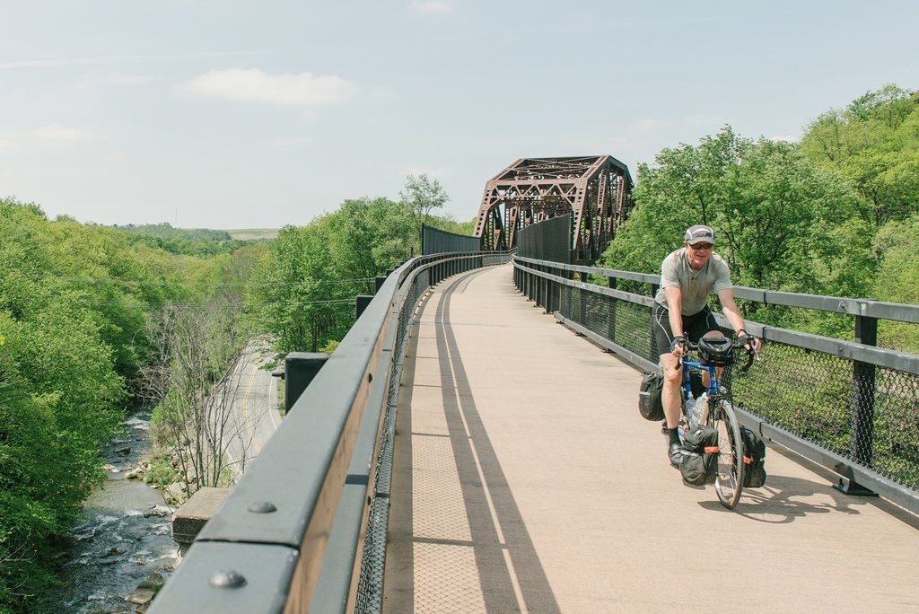 Cyclist rides along a raised bike path, bordered by lush green trees and crossing a metal bridge amid a natural landscape.