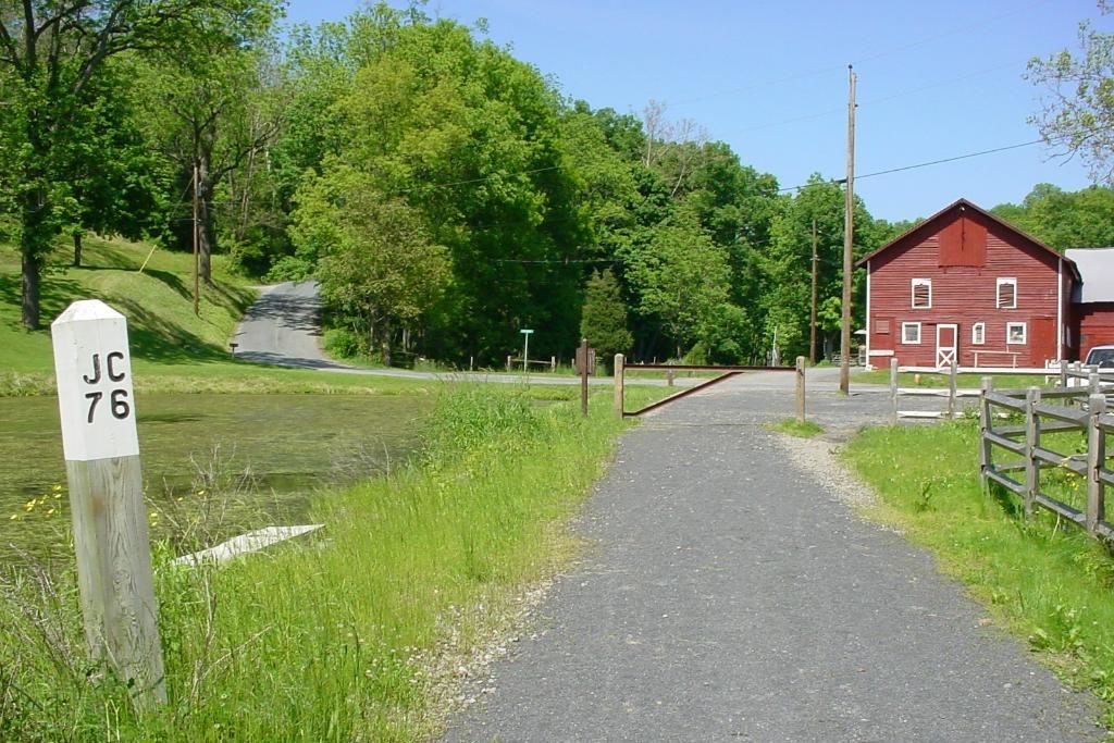 A gravel path leads to a red barn surrounded by lush greenery. A roadside post reads "JC 76" beside a grassy area.