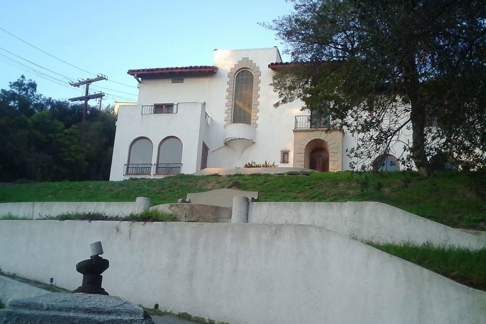 White stucco house stands on a grassy hill, surrounded by trees, with arched windows and a tiled roof under a clear sky.