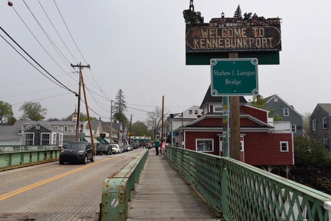 A bridge extends over a small-town street with houses and a "Welcome to Kennebunkport" sign. Cars and pedestrians are visible.