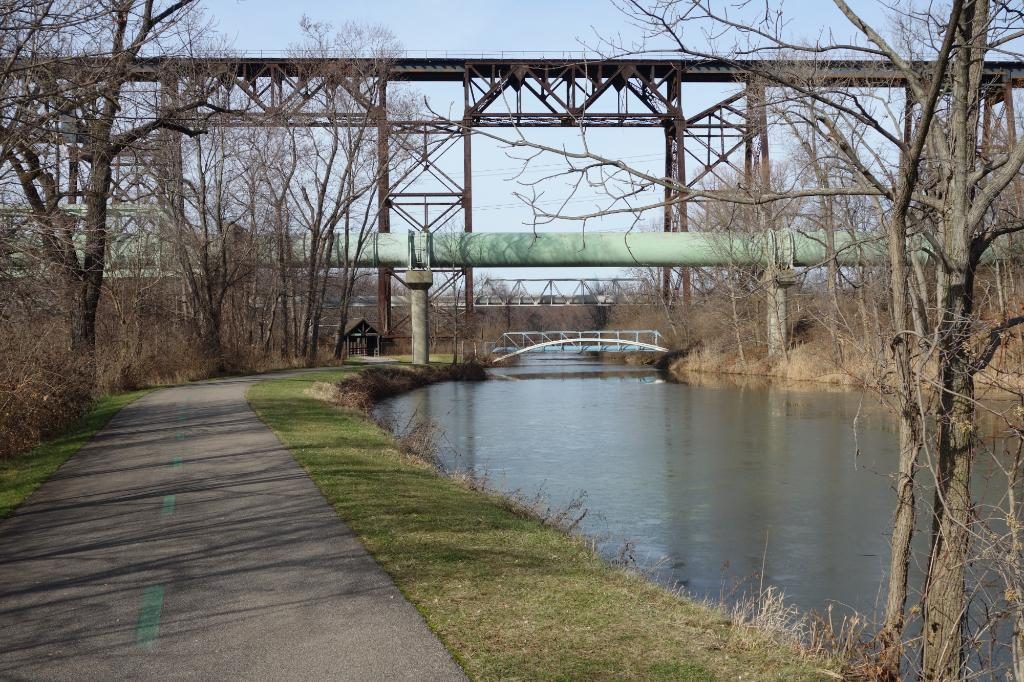 Path winds alongside a calm river, bordered by bare trees, with a large pipeline and metal bridge overhead in a serene outdoor setting.
