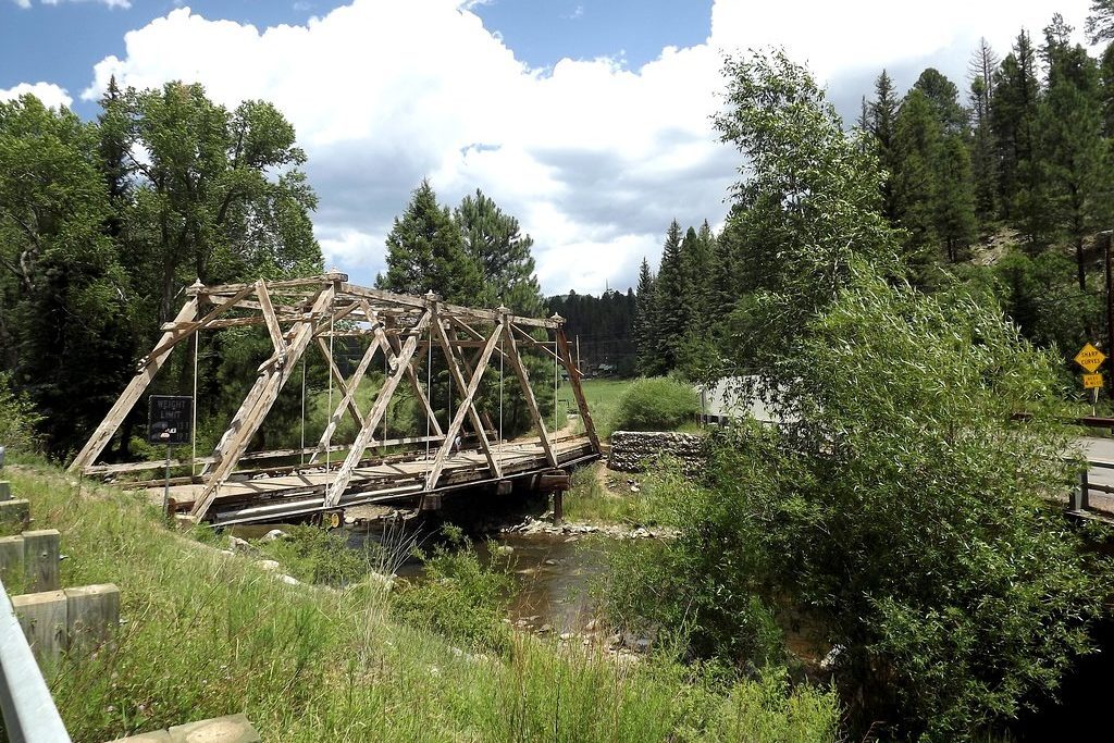 Wooden bridge stands over a small stream, surrounded by lush green trees and grass under a partly cloudy sky. Signs indicate weight limits.