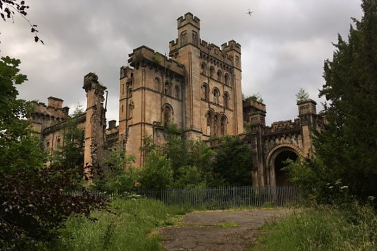 Castle stands silently, surrounded by overgrown greenery and cloudy sky, while an airplane flies above.