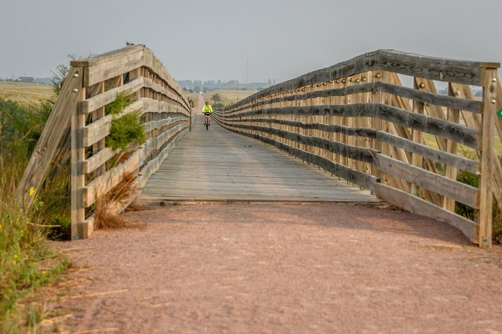 Cyclist rides along a wooden bridge; surrounded by open fields and distant horizon under a cloudy sky.
