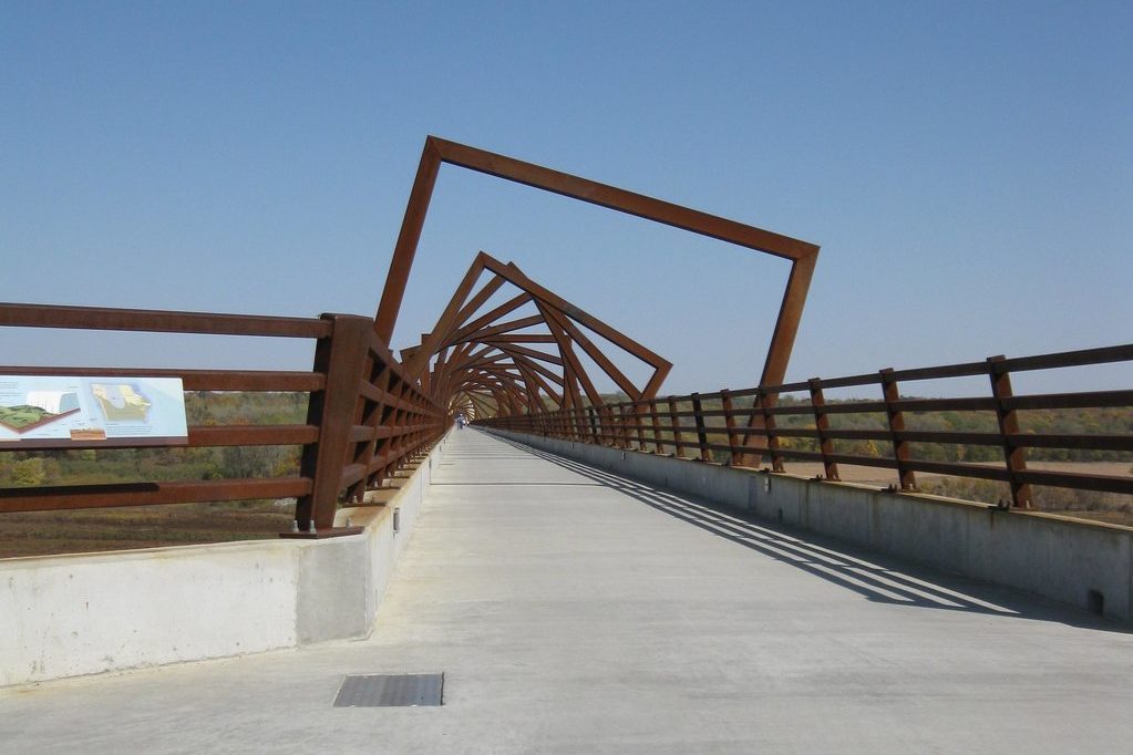 Rusty geometric bridge stretches across a landscape, featuring steel square frames. Sparse foliage and open sky surround the structure. Nearby signage includes text.