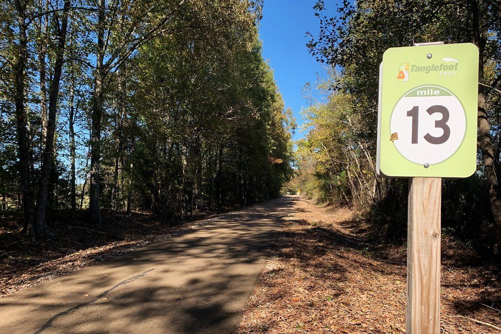 Trail sign displays "Tanglefoot Trail, mile 13" alongside a tree-lined path with dappled sunlight.