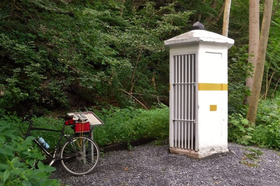 Bicycle rests beside a small, white, barred structure surrounded by dense greenery in a forested area.