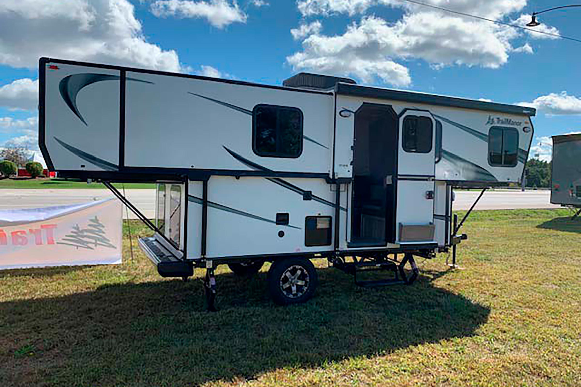 Camper trailer parked, door open, on grassy field under blue sky with clouds; nearby banner with tree symbols and partial text visible.