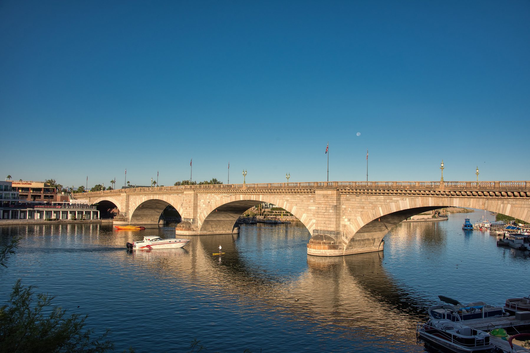 Bridge spans river; boats glide beneath; urban buildings and clear sky serve as backdrop.