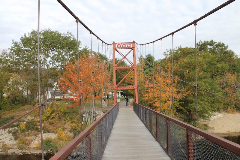 A suspension bridge extends toward orange-leaved trees, with a person walking in an autumn park setting.
