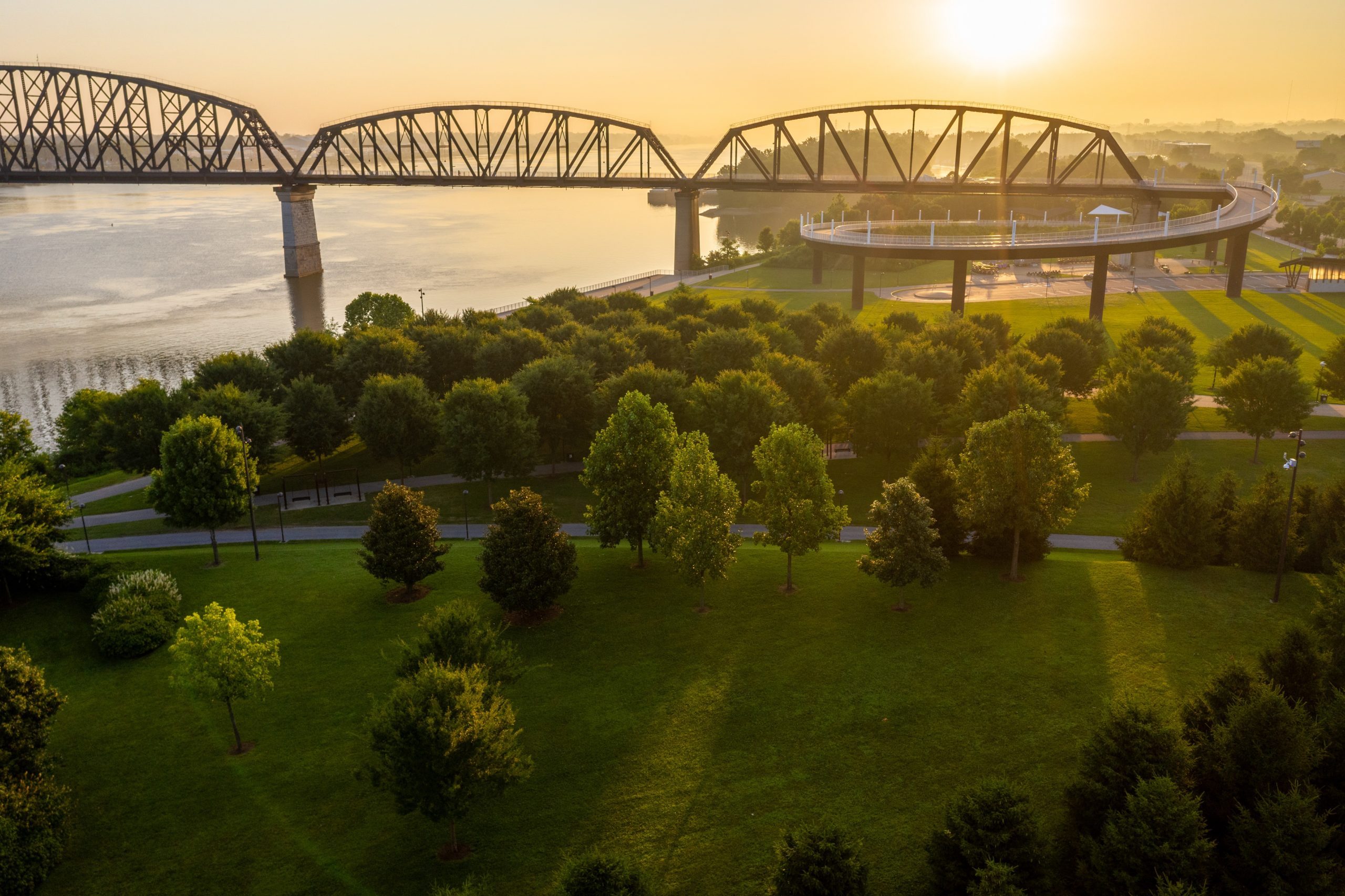 Bridge extends over a calm river, with a park filled with trees and paths underneath, bathed in warm sunset light.