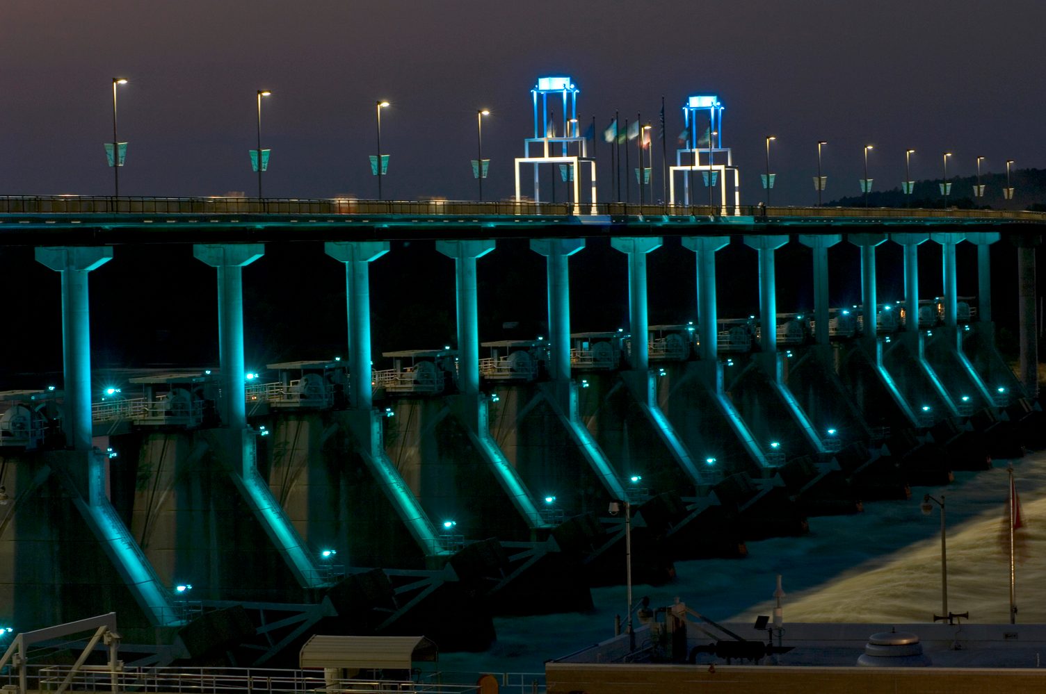A dam illuminated with blue lights spans a waterway at night, with streetlights and flags lining the top.