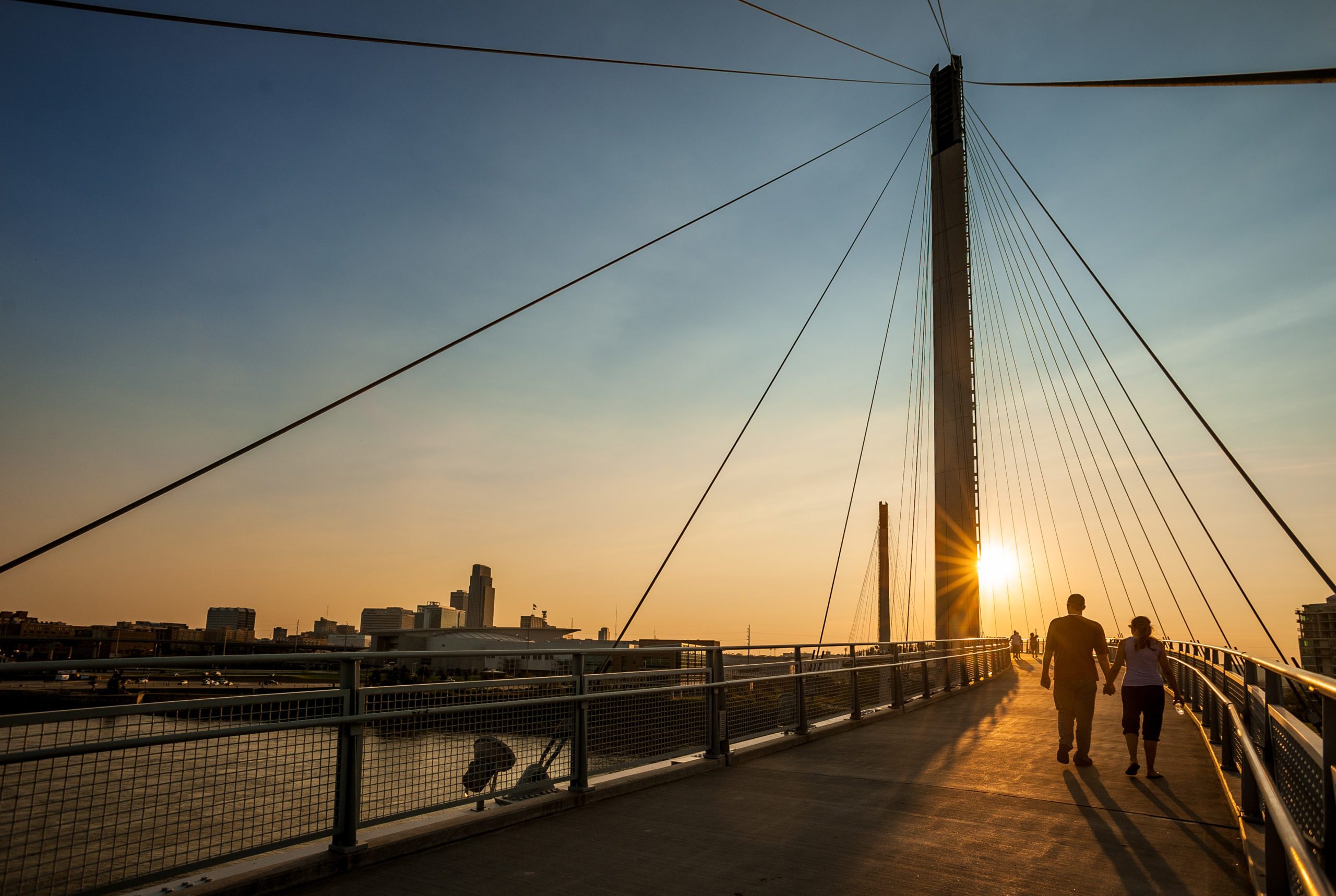 People walk across a suspension bridge at sunset, with city skyline silhouetted against the colorful sky.