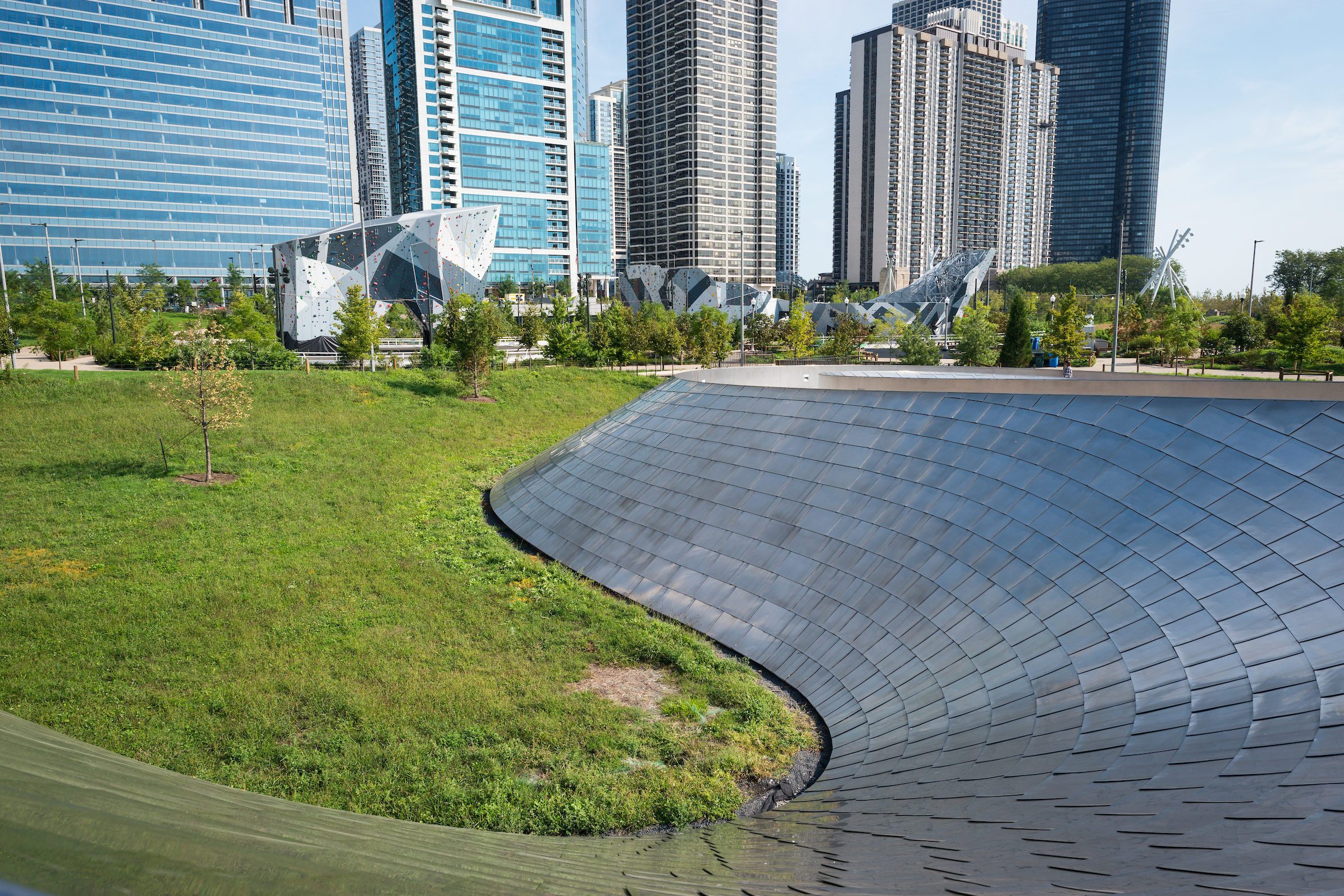 Curved metallic structure arcs beside a grassy area with modern skyscrapers in the background, amidst urban parklands.